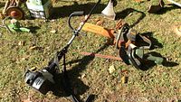 Full view of all three tools laid on the grass: Ryobi trimmer, Husqvarna chain saw with orange blade guard, Black & Decker hedge trimmer.