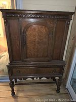 Full view of antique cabinet showing front with arched door, drawer, legs, and carved details