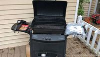 Char-Broil black gas grill with open lid, showing cooking grates and warming rack, side shelves holding grill tools and seasoning packets, propane tanks at base.