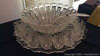 Large crystal punch bowl positioned on matching large crystal tray with scalloped edge, all with intricate cut-glass design, with 12 matching crystal cups arranged around the bowl on the tray.