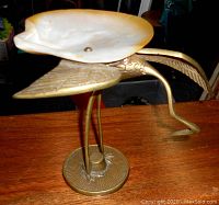 Front and side angle of vintage brass stork or crane statue with attached abalone shell dish on its back set on a wooden surface.
