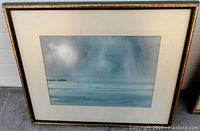 Framed watercolour artwork showing moody blue-grey landscape with sun or moon behind clouds, signed by Gordon Peters.