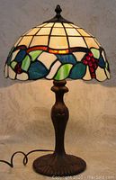 Full view of the stained glass lamp illuminated, showing the colorful floral design around the shade with blue and green leaves and red berries, cream-white glass panels, dark bronze base with finial top.