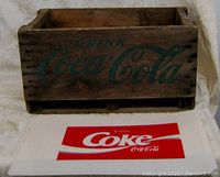 Wooden crate and flat plastic Coca Cola sign laid out side by side on a table for scale comparison.