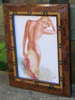 Framed burled wood print of a nude woman holding a large hat outdoors, side view showing frame and art clearly.