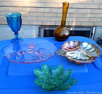 Five pieces including amber vase, blue goblet, pink bowl, green leaf dish, and shell-shaped dish on a blue table against a brick wall.