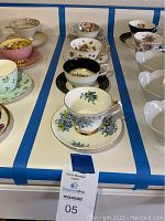 The photo shows 7 bone china tea cups arranged with matching saucers in various floral designs, placed on a white surface with blue tape lines.