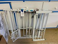 White metal pet gate and extension panel leaning against drawers, showing locking mechanism and gate door configuration.