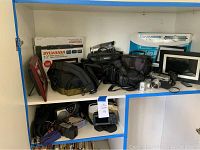 Shelf with digital photo frames, Panasonic video cameras, camera bags, Canon camera, CD player, and Garmin GPS.