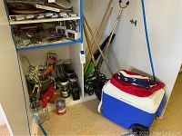 Wide view of cooler, various garden tools like rakes and shovels, metal and glass lanterns, cords, and flag folded on cooler.