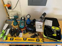 Photo of tools and accessories on a wooden surface including grills tools with green and black handles, two cordless screwdrivers (black and red), teal cordless drill and flashlight, battery chargers, and a yellow torpedo level.