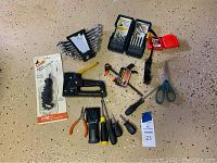 Image showing assortment of hand tools including screwdrivers, staple gun, wrenches, drill bits, measuring tapes, clamp, pliers, and scissors on a beige speckled floor.