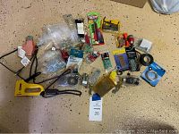 Wide shot showing assortment of tools and hardware on floor including Stanley staple gun, manual tire gauge, packs of drill bits, various screws, nails and washers, picture wire, and other hardware items.
