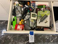 Top view of open drawer showing assorted electronic and utility items including battery charger, USB hub, cables, timer, scissors, batteries, and fire starters