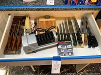 Drawer containing Iona electric knife, wooden-handled steak knives in tray, multiple black-handled chef knives, a knife sharpener, and some wooden cutting boards.