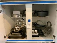 Photo of open cabinet shelf showing assorted metal baking pans, mixing bowls, bear-shaped mold, muffin tin, wire basket.