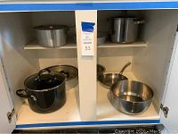 Various stock pots, mixing bowl, frypan and oversized cake pans arranged in a cabinet.
