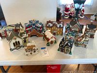 Overview photo of 11 assorted Christmas village houses and buildings displayed on a table, including a church with tall steeple and various smaller houses and shops.