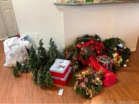 Photo showing several small artificial pine trees, decorated Christmas wreaths, garlands, and holiday storage boxes on the floor.