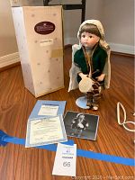 Full view of the doll standing on white stand with original box behind it and certificate of authenticity and photo card on the floor in front.