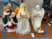 Photo showing three Christmas figures: animated girl with skates holding a candle, painted plaster angel with lit candle, and plastic Baby Jesus figure lying down.