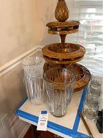 Wooden three-tier serving tower with carved edges and a wooden pineapple finial on top, accompanied by four glass vases of varying styles and heights on a white table.