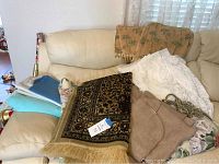 View of palm tree theme beige drapes folded on couch along with beaded valance, palm tree patterned beige blanket, multicolored comforter or throw, fabric remnants, and decorative fringe rug.