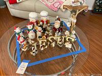Wide view of figurines on round glass table showing two nutcrackers, multiple snowmen, Santa Claus, crucifix, and wooden letters spelling JESUS