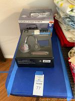 Two boxed items on a blue surface beside folded bedding - the front box is Sharper Image wireless headphones, back box is Sylvania portable CD radio boombox