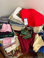 Photo showing stack of assorted tablecloths, placemats, and napkins including red crocheted placemats and various folded fabrics.