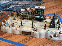 Front view of the animated Christmas village showing buildings, lampposts with string lights, dancing girl figurine, sleigh, and snow-covered base.