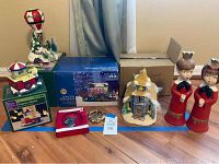 Photo showing multiple Christmas village decorations, boxed items and candlesticks on a wooden floor near a wall with curtain.