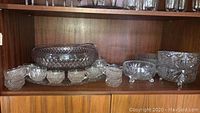 Wide view showing large glass punch bowl, multiple small matching glass cups, and other glass bowls in the background on wooden shelf