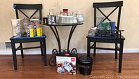 Wide shot showing all items including glassware on black chairs and floor, large glass vases, glass spice containers, spiral vegetable slicer box, Fry Daddy fryer, and chafing stand