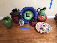 Wide view showing seven ceramic and porcelain pieces arranged on floor: tall green vase, two dark brown vases, rectangular striped ceramic bottle, large blue-green decorative bowl on metal stand, tall colorful pitcher, two decorative plates with fruit motifs.