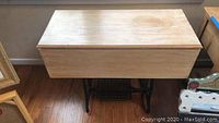 Drop leaf wooden table closed with worn, cracked light wood top showing grain. Mounted on black antique cast iron sewing machine base with treadle