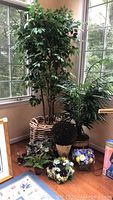 Photo of the lot showing a tall faux tree in a woven basket alongside a medium faux palm tree in a ceramic pot and various smaller faux plants and floral arrangements on the floor.