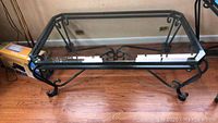 Full view of rectangular glass top coffee table with black metal scrollwork frame and curved legs on hardwood floor.