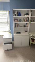 Photo of tall white cabinet with three open shelves filled with books and decor, and two lower shelves behind closed doors, some contents inside; located against blue wall next to window with blinds