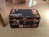 Top and front view of black steamer trunk with multiple political and music stickers and metal trim.