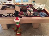 Wide view showing Longaberger basket liners, kitchen towels, Christmas glasses, assorted linens, and boxes containing miscellaneous decorative items on billiards table.