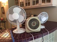 Three items on tiled counter: two white oscillating fans and a beige Vornado heater between them