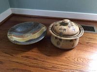 Photo of two pottery items placed on wooden floor; one is a bowl with wavy striped pattern, the other is a lidded pot with decorative markings.
