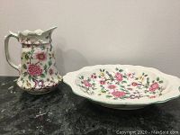 Photo showing jug and bowl side by side on a black surface against cream wall, displaying floral pattern.