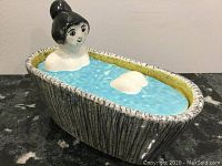 Front side view showing woman figurine emerging from blue water in bathtub shaped dish