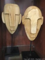 Two wood carved masks mounted on black metal stands, displayed in a cabinet.