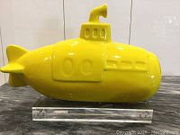 Side view of a glossy yellow submarine shaped ceramic bank on a clear acrylic stand showing raised details.