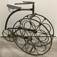 Front and side view of metal bicycle wine rack showing large front wheel with circular bottle holders and smaller rear wheel, seat and handlebars visible.