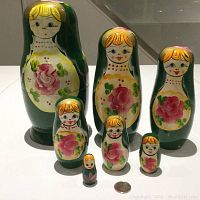 Seven Russian Matryoshka stacking dolls arranged in a row from largest to smallest with a coin for scale, showing detailed handpainted design with green base and pink roses.