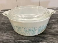 White opaque Pyrex glass bowl with light blue farm-themed pattern around the exterior, shown with clear glass lid on top on a dark surface.